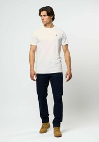 Vyras dėvi baltą trumpomis rankovėmis marškinėlį, tamsius džinsus ir rudas darbo batus, stovėdamas priešais paprastą baltą foną.