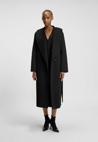 Long black coat with a broad collar, two buttons, and a straight cut. Worn over a black dress, complemented by high-heeled ankle boots.