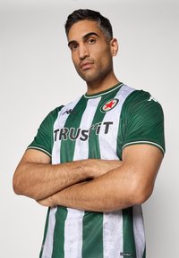 Mann med foldede armer iført en grønn og hvitstripet fotballtrøye med "TRUSTIT"-logo og et rødt stjernesymbol på brystet.