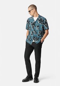 T-shirt met korte mouwen in groenblauw met een zwart bloemmotief, gecombineerd met zwarte slim-fit broek en zwarte schoenen. Model staat met handen in de zakken.