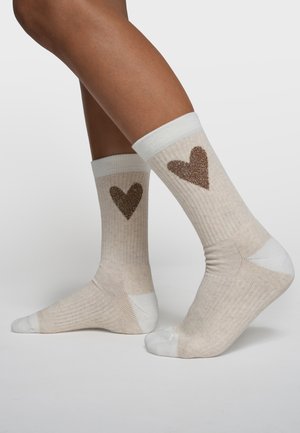 Beine tragen beige gerippte Socken mit weißen Zehen und Fersen, jeweils mit einem großen braunen Glitzerherz an der Außenseite.