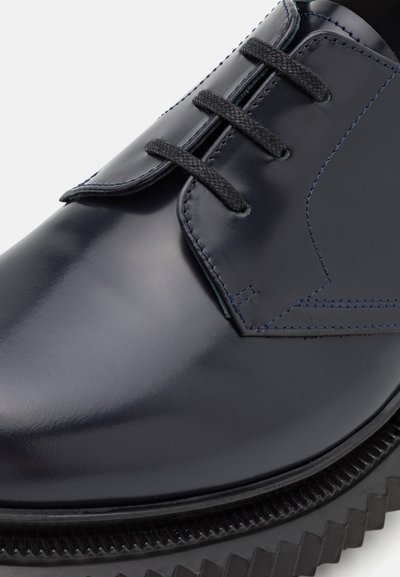 Pantofii din piele bleumarin, cu un finisaj neted, vârful rotund și șireturi negre. Prezintă cusături albastre contrastante și o talpă neagră zgâriată.