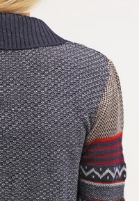 Sweter dzianinowy z teksturowanym granatowym korpusem, ribbować kołnierzem oraz wzorzystymi rękawami w kolorze beżowym, czerwonym i szarym z akcentami geometrycznymi.