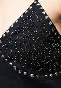 Μαύρο ύφασμα με ασημένια στρογγυλά τρουκς τοποθετημένα σε αφηρημένα, κυματιστά μοτίβα, περιβαλλόμενα από μεγαλύτερα ανακλαστικά τρουκς κατά μήκος της άκρης.