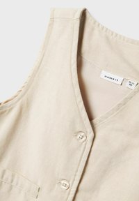 Helles beige ärmelloses Oberteil mit V-Ausschnitt, das zwei Knöpfe auf der Vorderseite und eine einzelne Brusttasche hat. Aus weichem Stoff gefertigt.