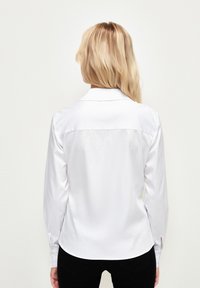 Chemise blanche à manches longues avec un col ajusté, une texture de tissu lisse et des boutons sur les poignets, vue de dos.