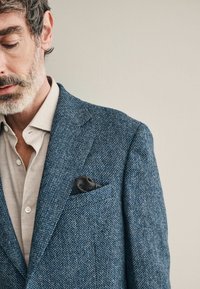 Plavi tweed sako s teksturiranim uzorkom, s crnom maramicom u džepu i svjetlosmešavom košuljom s kragnom ispod.