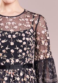 Robe noire avec une superposition transparente brodée de motifs floraux roses. Manches évasées avec des accents en perles, ruban en soie à la taille.