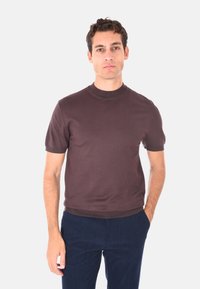 Uomo con capelli ricci e scuri, che indossa un maglione marrone a maniche corte e pantaloni blu navy, in piedi con una mano in tasca su uno sfondo bianco.