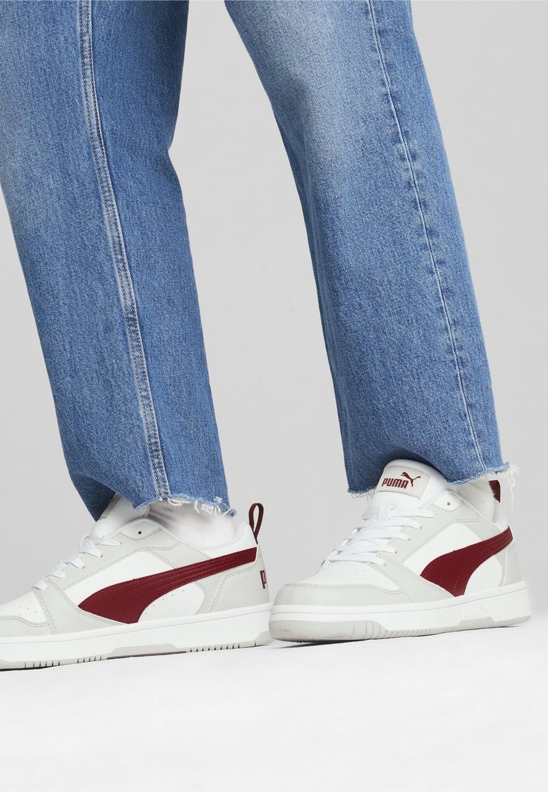 Personne portant un jean bleu clair à ourlets effilochés et des baskets Puma blanches avec des logos rouges, debout sur un fond uni clair.