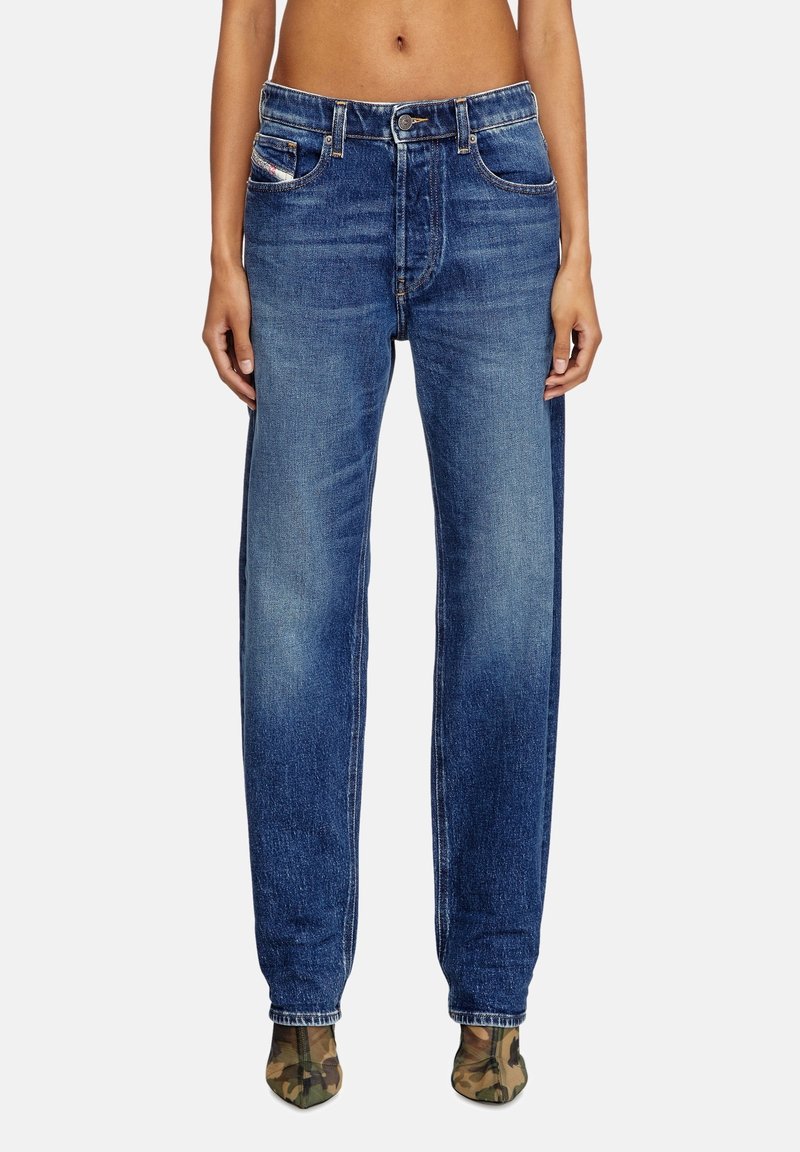 Personne portant un jean bleu taille haute et des bottes camouflage à bout pointu, debout devant un fond clair uni.