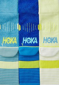 Three pairs of HOKA socks: light blue, dark blue with stripes, and yellow with stripes. Knit design with textured patterns throughout.