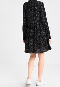 Robe noire à pois blancs, manches longues et jupe évasée. Confectionnée en tissu léger, finition avec un bouton au niveau du poignet.