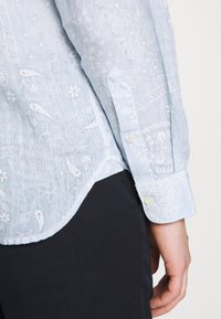 Jasnoniebieska koszula z teksturowanej tkaniny, ozdobiona białymi wzorami kwiatowymi i paisley. Posiada zapięcie na guziki przy mankietach oraz zaokrąglony dół.