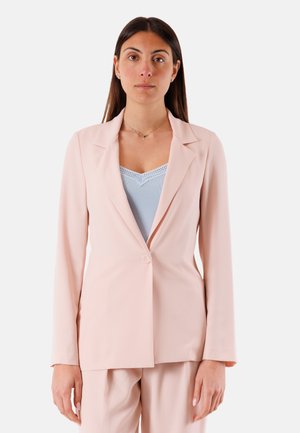 Donna che indossa un blazer rosa pallido e pantaloni coordinati su una maglia azzurra con bordo in pizzo, in piedi davanti a uno sfondo semplice.