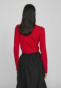 Röd randig långärmad topp med axelknappar, ihopparad med en svart kjol med elastisk midja. Tyget verkar mjukt med en figurnära silhuett.