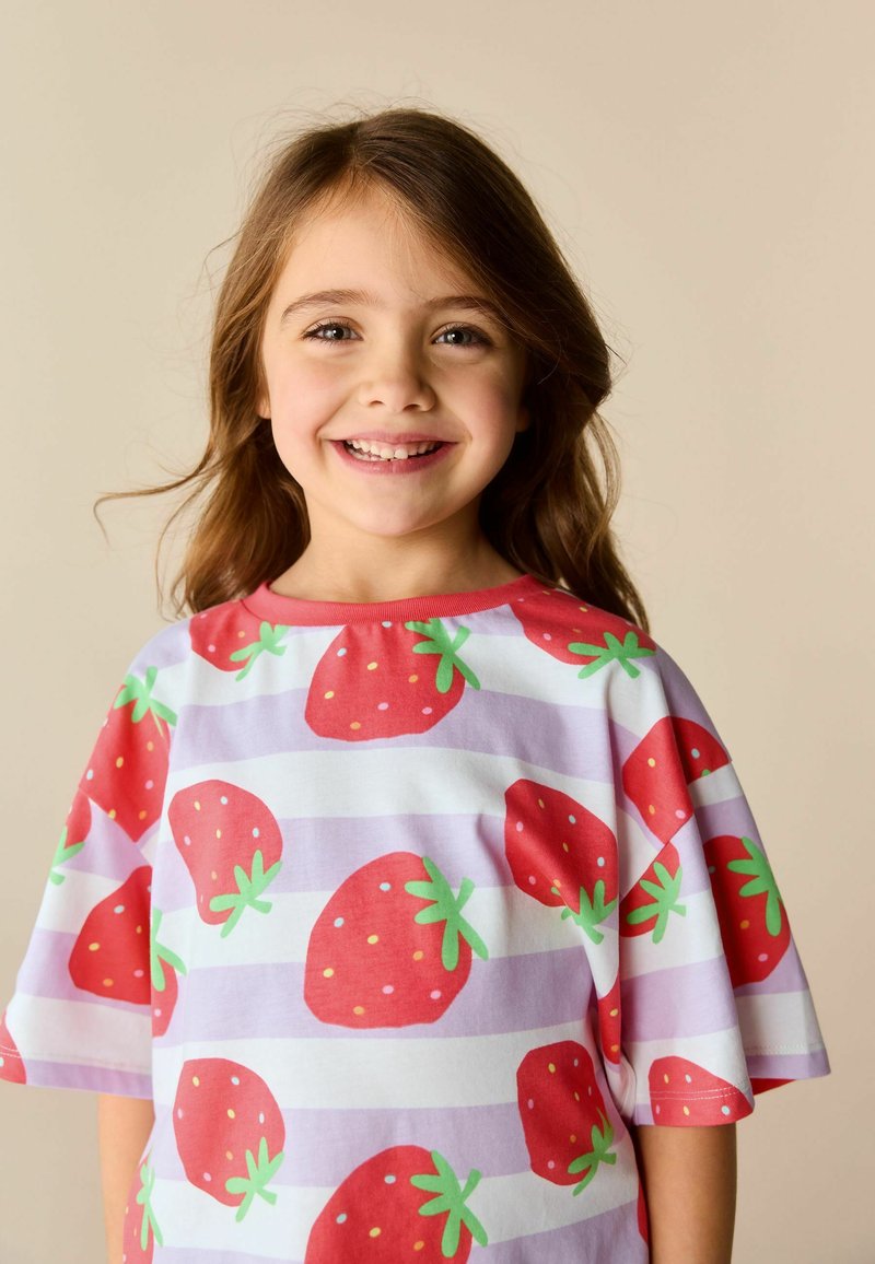 Joven sonriente con cabello largo y castaño, que lleva una camiseta a rayas blancas y lavanda cubierta de grandes fresas rojas sobre un fondo beige.