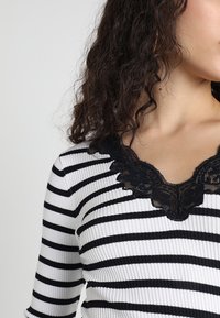 Haut à rayures noires et blanches en tissu côtelé, avec une dentelle bordant l'encolure en V et des manches trois-quarts.