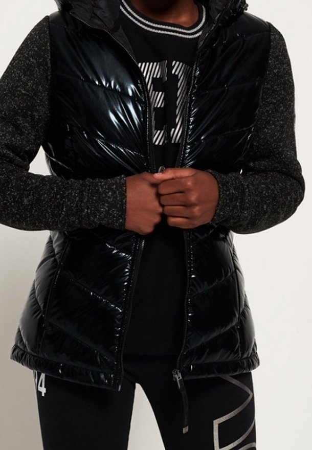 Personne portant une doudoune noire brillante sur un pull foncé, leggings noirs à motifs blancs, mains jointes à la taille, torse visible.