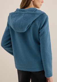 Veste en polaire bleue douce et texturée avec capuche, présentant une coupe décontractée et un design sans coutures, vue de dos.