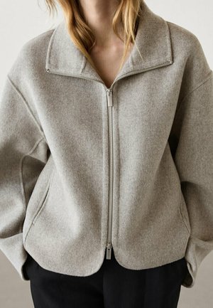 Femme portant une veste en laine gris clair zippée avec un large col et un pantalon noir, debout devant un fond neutre.