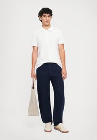 Weißes Baumwoll-Polohemd, navyfarbene Leinenhose, helle Sneakers und eine beige Tragetasche. Einfaches Design mit lockerer Passform, glatter Textur und ohne Muster.