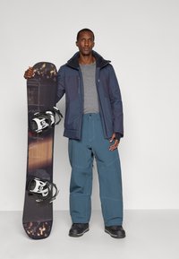 Čovjek u plavoj jakni i hlačama za snijeg drži uspravno snowboard s crnim i smeškim dizajnom protiv jednostavne pozadine.