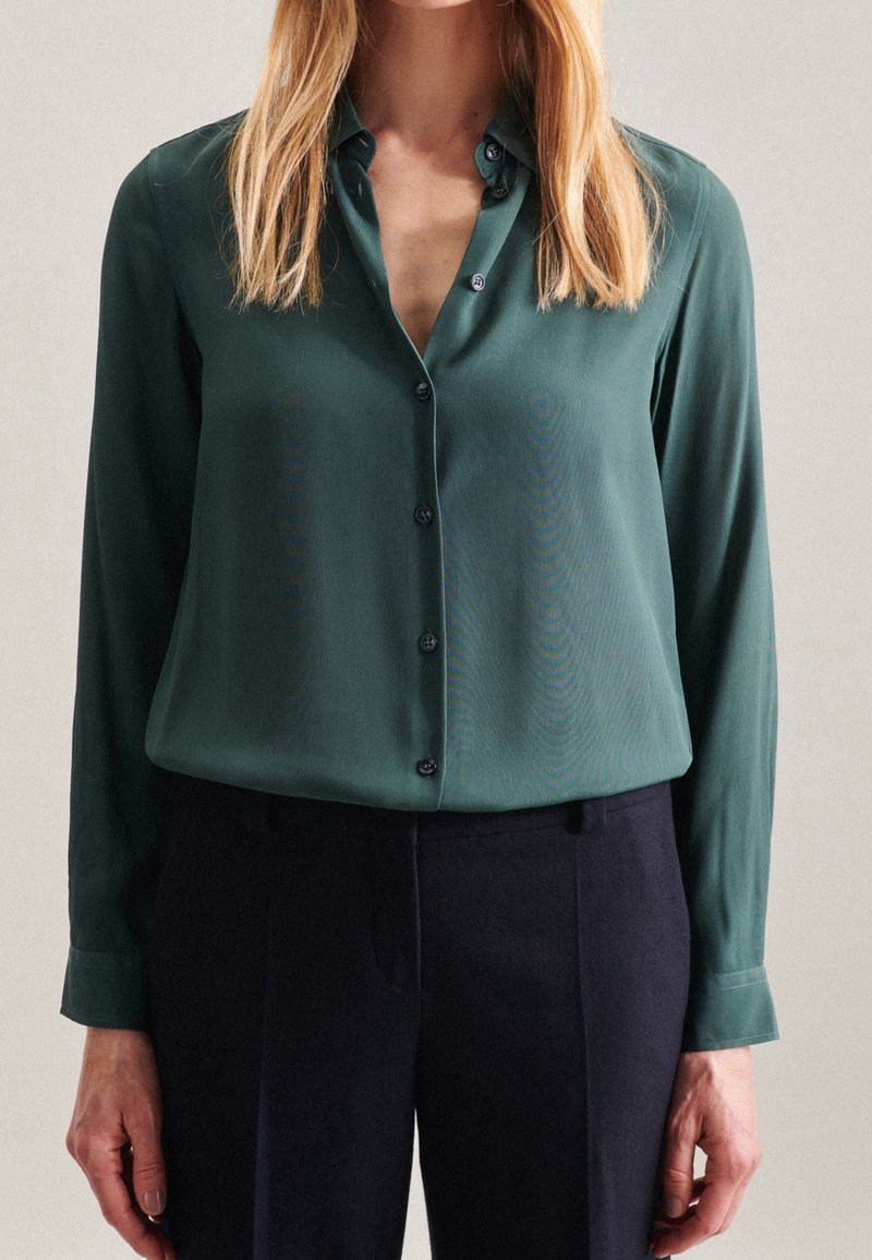 Donkergroen, blouse met lange mouwen en een kraag, met een knoopsluiting aan de voorkant en een gladde, lichte stof; gecombineerd met donkere broeken.