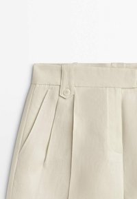 Pantalon beige taille haute avec passants de ceinture, patte boutonnée, poches latérales et plis sur le devant, fabriqué en tissu texturé.