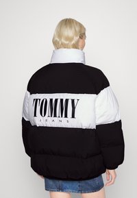 Giubbotto imbottito bianco e nero con collo alto, vestibilità oversize e grande logo "TOMMY JEANS" sulla schiena. Morbido, con texture trapuntata.