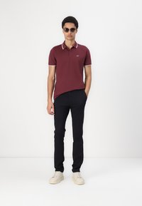 Maroon polo shirt with white collar accents, paired with black trousers and white sneakers. Model wearing sunglasses stands against a plain background.