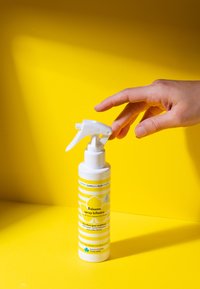 Condizionatore spray bifasico in una bottiglia cilindrica bianca con strisce gialle, dotato di uno spruzzatore bianco. Lo sfondo è di un giallo brillante.