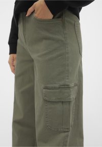 Khaki cargo kalhoty vyrobené z bavlny, které mají boční kapsy, uvolněný střih a šité švy, s hladkou texturou a minimálními detaily.