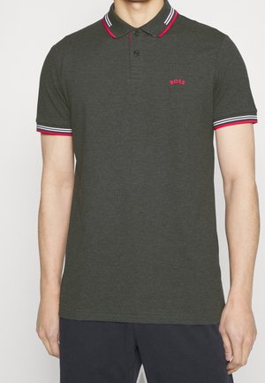 Mann trägt ein dunkelgraues Poloshirt mit rotem und weißem Abschluss an Kragen und Ärmeln, rotes „BOSS“-Logo auf der linken Brust, Hände an den Seiten.