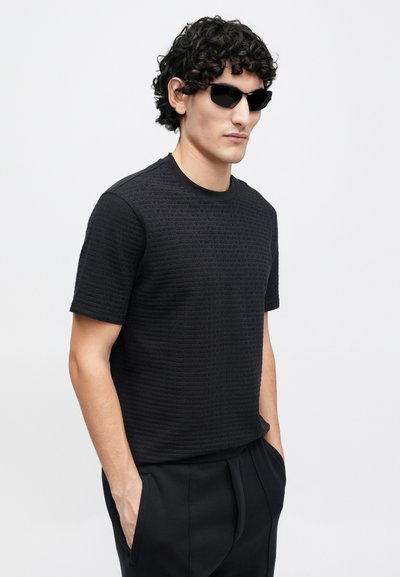 Čierne tričko s krátkym rukávom z textúrovaného materiálu s jemným vzorom, doplnené tmavými slnečnými okuliarmi a čiernymi nohavicami.