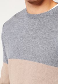 Pull en maille gris et beige avec un col rond et un bloc de couleur horizontal, présentant une texture lisse et un tissu léger.