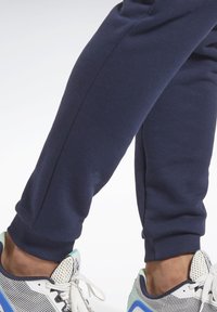 Mørkeblå sweatpants med ribbede manchetter, lavet af blødt materiale. En hvid sneakers med mesh-detaljer og blå-grønne accenter.