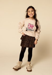 Jong meisje dat glimlacht met lang haar, gekleed in een beige sweatshirt met een hartontwerp, een rok met luipaardprint, zwarte panty’s en beige laarzen.