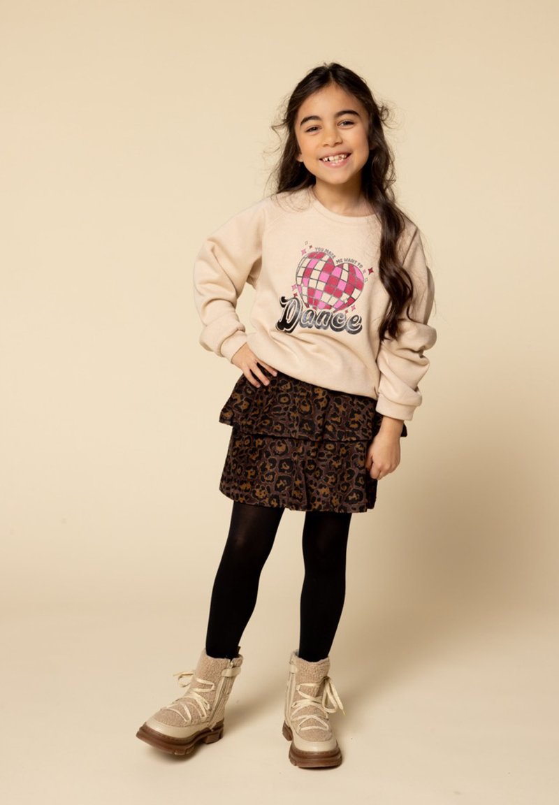 Jong meisje dat glimlacht met lang haar, gekleed in een beige sweatshirt met een hartontwerp, een rok met luipaardprint, zwarte panty’s en beige laarzen.