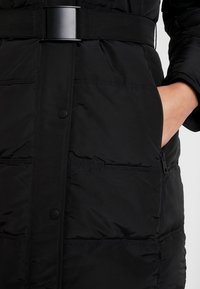 Abrigo acolchado negro con cintura ajustable, botones a presión y bolsillos laterales visibles. La tela tiene una textura suave con segmentos acolchados.
