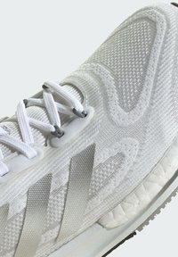 Weiße Sportschuhe mit einem strukturierten Mesh-Obermaterial, silbernem Drei-Streifen-Design und einer gepolsterten Sohle mit einem gemusterten Profil.