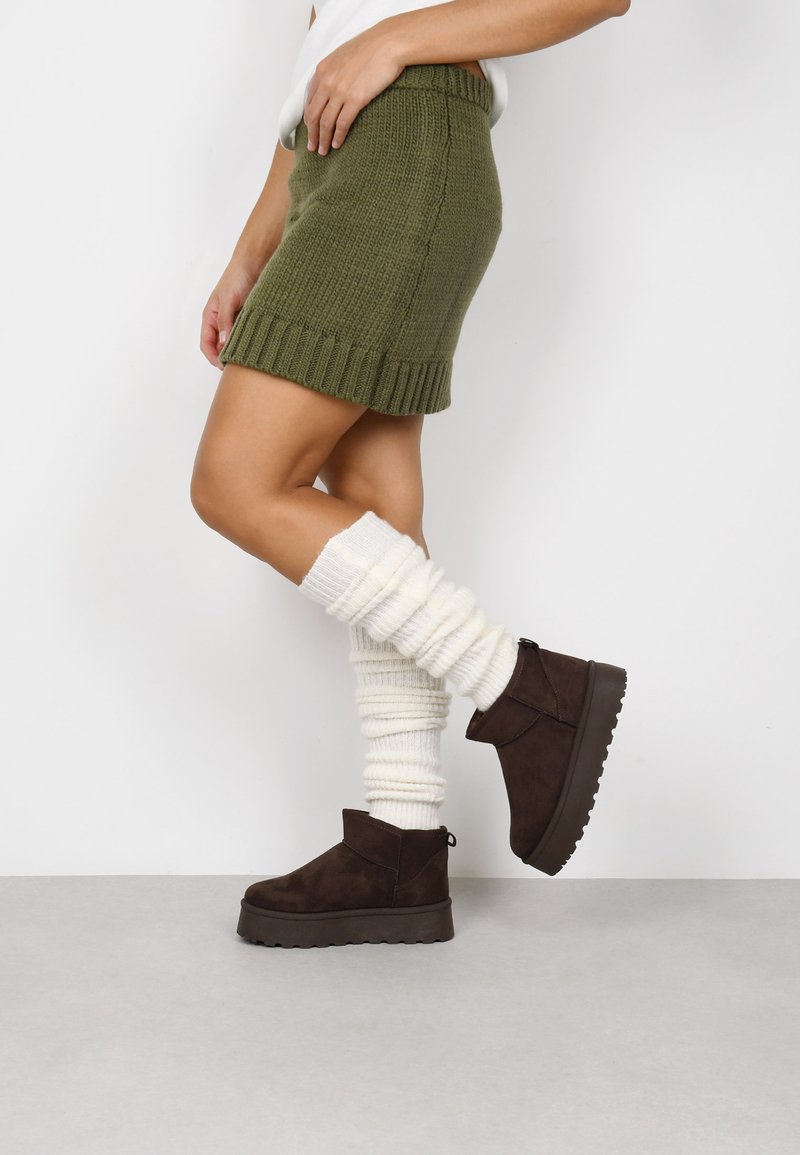 Falda mini de punto verde oliva con dobladillo acanalado, combinada con calentadores de piernas de punto color crema y botas de tobillo marrones con superficie texturizada.