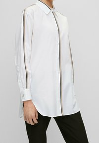 Bílá košile s dlouhými rukávy a černými a hnědými bočními pruhy, s límečkem na knoflíky a manžetami. Hladký povrch a přiléhavý střih.