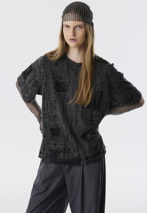 Chemise noire oversize avec un imprimé texturé, dotée de manches courtes et de détails en maille aux poignets. Portée avec un pantalon gris ample et un bandeau en maille.