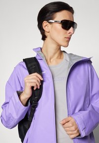 Jasnofioletowa wodoodporna kurtka z szarym wnętrzem, czarnym paskiem plecaka, szarym podkoszulkiem i dużymi czarnymi okularami przeciwsłonecznymi. Posiada zamki błyskawiczne i detale szwów.