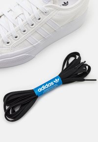 Λευκό Adidas sneaker με ανοιχτό γκρι ρίγες και μαύρα κορδόνια Adidas με μπλε ετικέτα σε λευκή επιφάνεια.
