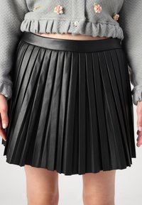 Jupe en cuir plissé noir avec une taille lisse, présentant des plis verticaux et un ourlet net. Le tissu a une finition polie et brillante.