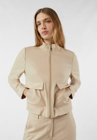 Beżowa kurtka na zamek z wysokim kołnierzem, dwoma przednimi kieszeniami z patkami oraz gładką, miękką fakturą. Stylizowana z dopasowanymi spodniami o minimalistycznym designie.