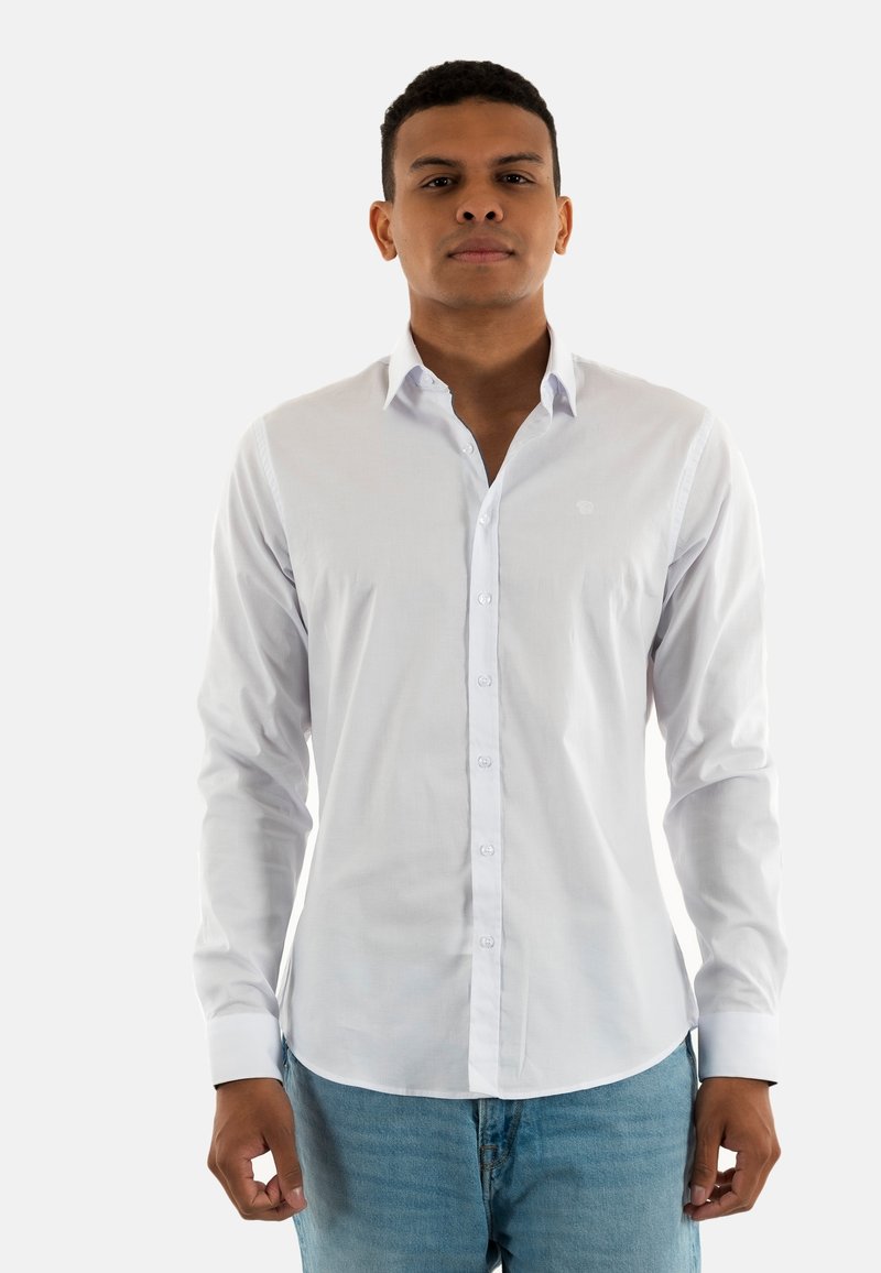 Chemise blanche à manches longues en tissu rigide, dotée d'une fermeture à boutons sur le devant et d'un petit logo sur la poitrine. Associée à un jean bleu clair.