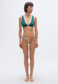 Γυναίκα που στέκεται ξυπόλυτη με πράσινο τοπ μπικίνι και πολύχρωμα μαγιό με κορδόνια στο πλάι, κοιτάζει προς τα δεξιά μπροστά σε ένα απλό φωτεινό φόντο.